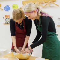 赤いエプロンと緑のエプロンの女性2人が、ボウルの中で生地作り(粉と水で生地を作る工程)をしている 手打ちうどん作り体験教室 2025年12月 A6407348-1.jpg – イリコスキー製麺所