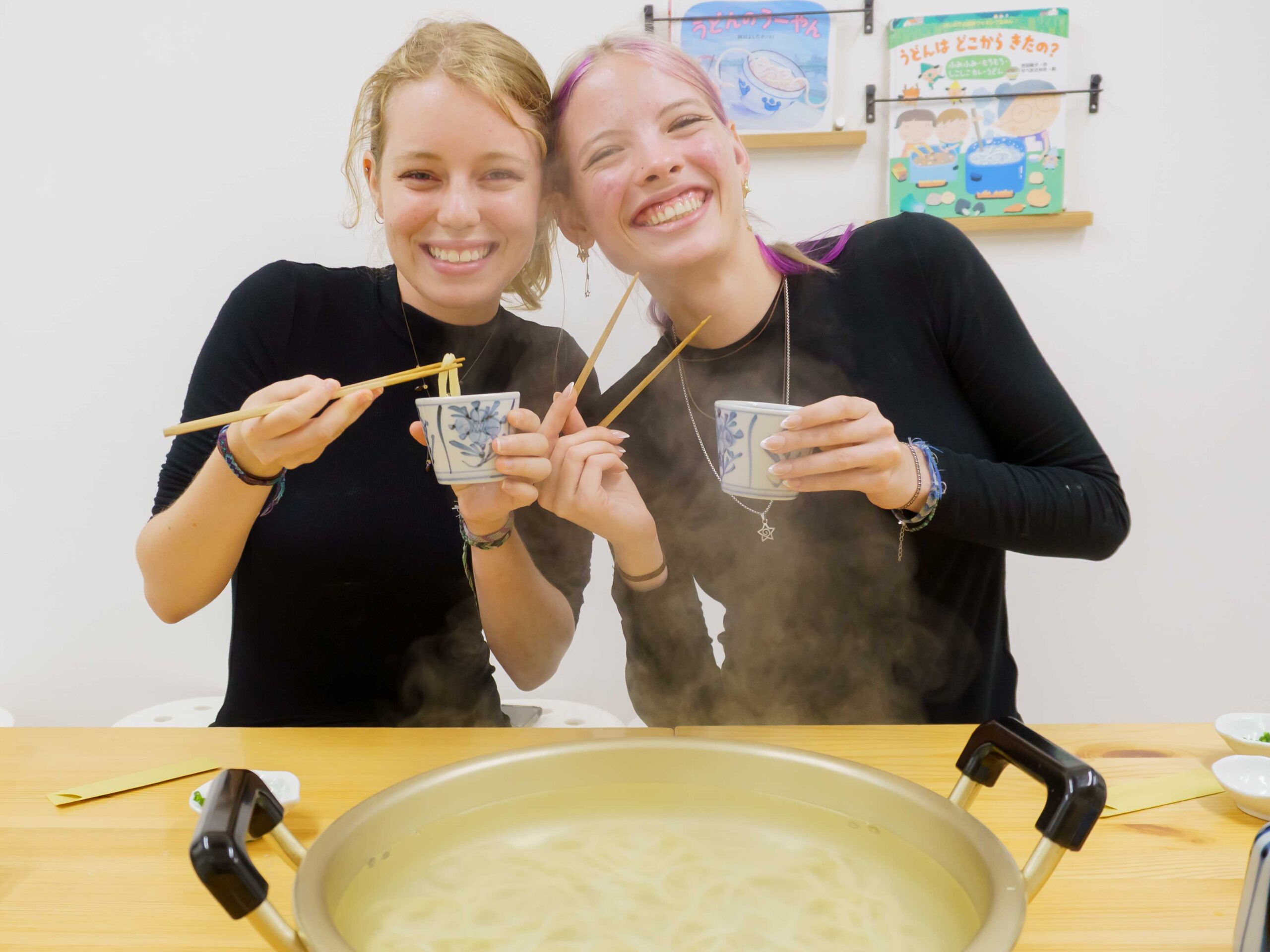 女性2人が湯気の立つ鍋の前で、箸と蕎麦猪口を持ってうどんを味わっている 手打ちうどん作り体験教室 2025年12月 A6407372-1-scaled.jpg – イリコスキー製麺所
