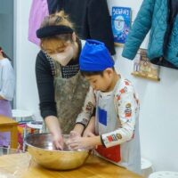 ビニール袋に入れたうどん生地を丁寧にまとめる女性と見守る女の子 / A woman carefully forming udon dough inside a plastic bag while a young girl watches. 手打ちうどん作り体験教室 2025年12月 A6407661-1.jpg – イリコスキー製麺所