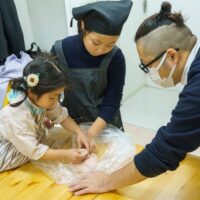 テーブルの上でうどん生地を一生懸命にまとめるお子様たちと見守る男性 / Children working hard to form udon dough on a table while a man watches. 手打ちうどん作り体験教室 2025年12月 A6407677-1.jpg – イリコスキー製麺所