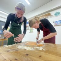 緑のエプロンの女性が切ったうどんを持ち上げ、赤いエプロンの女性が麺を切っている 手打ちうどん作り体験教室 2025年12月 IMG_1074.jpg – イリコスキー製麺所