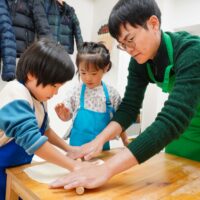 男性が子供たちの手元に添えて一緒に麺棒で生地を薄く伸ばしている様子。 / A man helping children roll out the dough thinly with a rolling pin. 手打ちうどん作り体験教室 2025年12月 A640733-1-1.jpg – イリコスキー製麺所