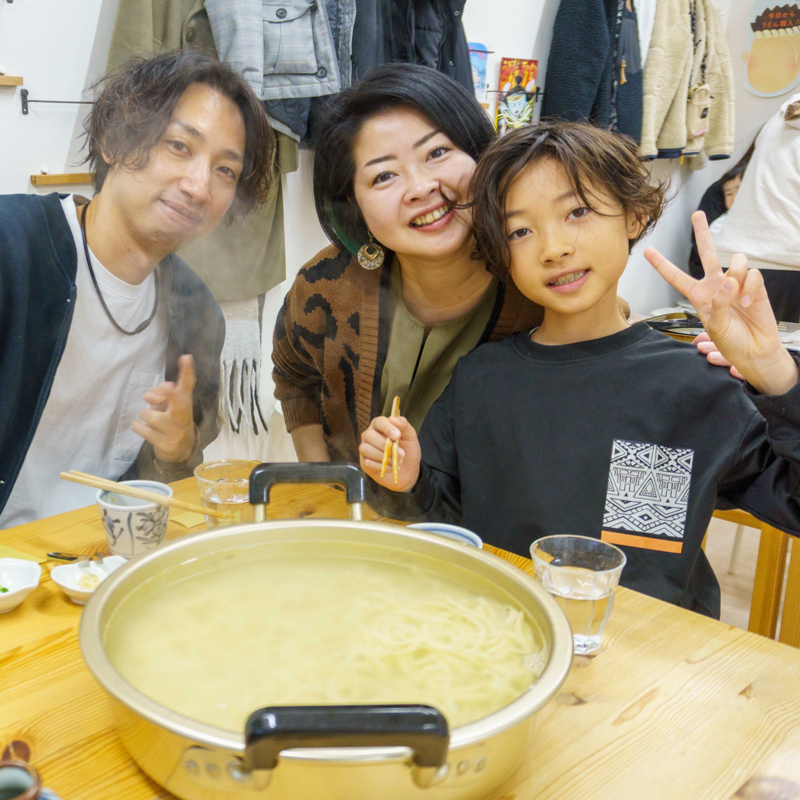 茹でたてのうどんが入った大きな鍋を囲んで笑顔を見せる家族。Steam rising from a large pot of fresh udon surrounded by a smiling family. 手打ちうどん作り体験教室 2026年1月 A6407773-1-scaled.jpg – イリコスキー製麺所