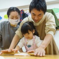 麺棒をしっかりと握り、体重をかけてピンク色の生地を伸ばす女の子 / A girl firmly grasping a rolling pin and putting her weight into stretching the pink dough. 手打ちうどん作り体験教室 2026年1月 A6407825-1.jpg – イリコスキー製麺所