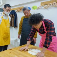 ピンクのエプロン姿で生地を伸ばす様子を真剣に見つめる男性ゲストたち / Male guests watching intently as a man in a pink apron rolls out the dough. 手打ちうどん作り体験教室 2026年1月 A6407912-1.jpg – イリコスキー製麺所