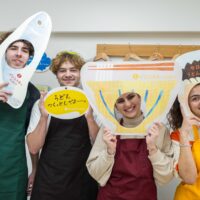 うどんや魚のイラストが描かれた顔はめパネルを持って楽しむ4人の若者 Four young people having fun holding comic foregrounds (kao-hame panels) with illustrations of udon and fish 手打ちうどん作り体験教室 2026年1月 A6408146-1.jpg – イリコスキー製麺所