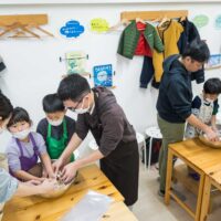 エプロンとマスクをつけ、ボウルの中で粉と水を混ぜてうどんの生地作りに真剣に取り組む2組の友人グループ alt: Two groups of friends wearing aprons and masks, seriously working on mixing flour and water in bowls to make udon dough. 手打ちうどん作り体験教室 2026年1月 A6408177-1.jpg – イリコスキー製麺所