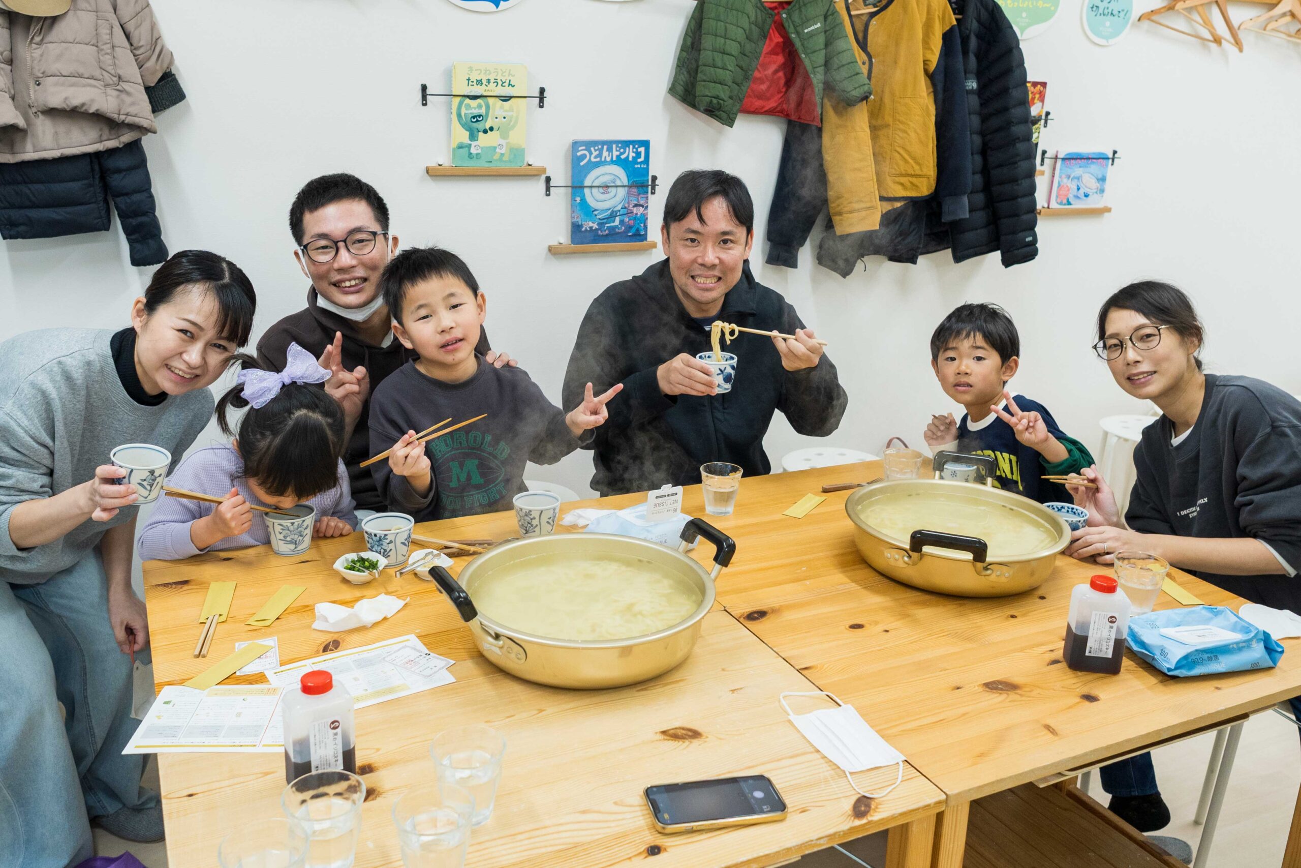 大きな鍋のうどんを囲み、箸やピースサインで笑顔を見せる2組の友人グループ alt: Two groups of friends smiling with chopsticks and peace signs around large pots of udon. 手打ちうどん作り体験教室 2026年1月 A6408189-1-scaled.jpg – イリコスキー製麺所