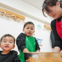粉と水を混ぜて生地作りをするお兄ちゃんと、それを見守るお母さんと弟。 (Older brother mixing flour and water to make dough, watched by his mother and younger brother.) 手打ちうどん作り体験教室 2026年1月 A6408220-2.jpg – イリコスキー製麺所