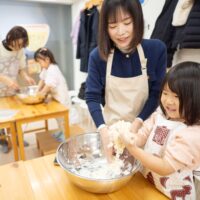 ボウルの中で小麦粉と水を混ぜて生地を作る女の子と、見守るお母さん。 Girl mixing flour and water in a bowl to make dough, with her mother watching. 手打ちうどん作り体験教室 2026年1月 A6408452-1.jpg – イリコスキー製麺所