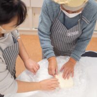 踏みの合間にうどん生地を折りたたんでいる2人の参加者。Two participants folding udon dough between stepping
rounds.