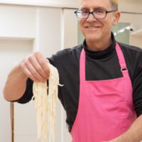 イリコスキー製麺所うどん作り体験教室で完成した手打ちうどんの生麺を高く掲げ、満面の笑みを見せる海外ゲスト。背景にイリコスキー製麺所のロゴとバナーが見える。An international guest at Iricosky Udon Making Experience Class lifting freshly made udon noodles high with a broad smile; the Iricosky logo and banner visible in the background. 手打ちうどん作り体験教室 2026年3月 iricosky-udon-class-fresh-noodles-lifted-smile-scaled.jpg – イリコスキー製麺所
