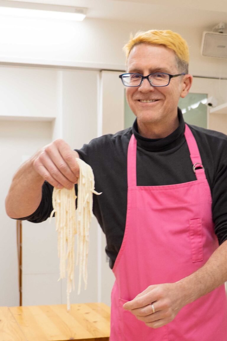 イリコスキー製麺所うどん作り体験教室で完成した手打ちうどんの生麺を高く掲げ、満面の笑みを見せる海外ゲスト。背景にイリコスキー製麺所のロゴとバナーが見える。