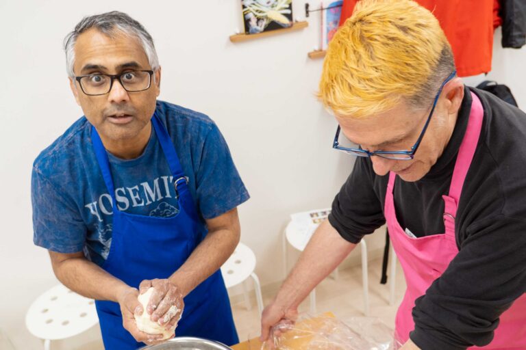 イリコスキーうどん作り体験教室で粉と水を混ぜた生地をひとかたまりにまとめている海外ゲスト2人。青エプロンの方がカメラに目を向けている。 手打うどん作り体験教室 2026年1月 udon-class-dough-gathering-two-guests-osaka.jpg – イリコスキー製麺所