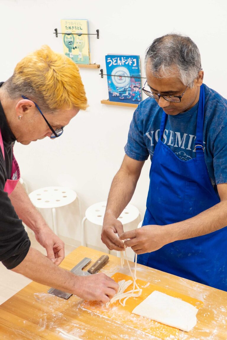 イリコスキーうどん作り体験教室で麺切りを終えた直後、2人の海外ゲストがテーブルの上で麺を引き出している場面。包丁とオレンジのまな板、打ち粉が並ぶ。 手打うどん作り体験教室 2026年1月 udon-class-noodle-cutting-two-guests-osaka.jpg – イリコスキー製麺所