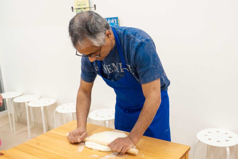 イリコスキーうどん作り体験教室で麺棒を両手で押さえながら生地を延ばす海外ゲスト。打ち粉の舞うテーブルで集中した横顔が印象的。 手打うどん作り体験教室 2026年1月 udon-class-rolling-dough-guest-profile-osaka.jpg – イリコスキー製麺所