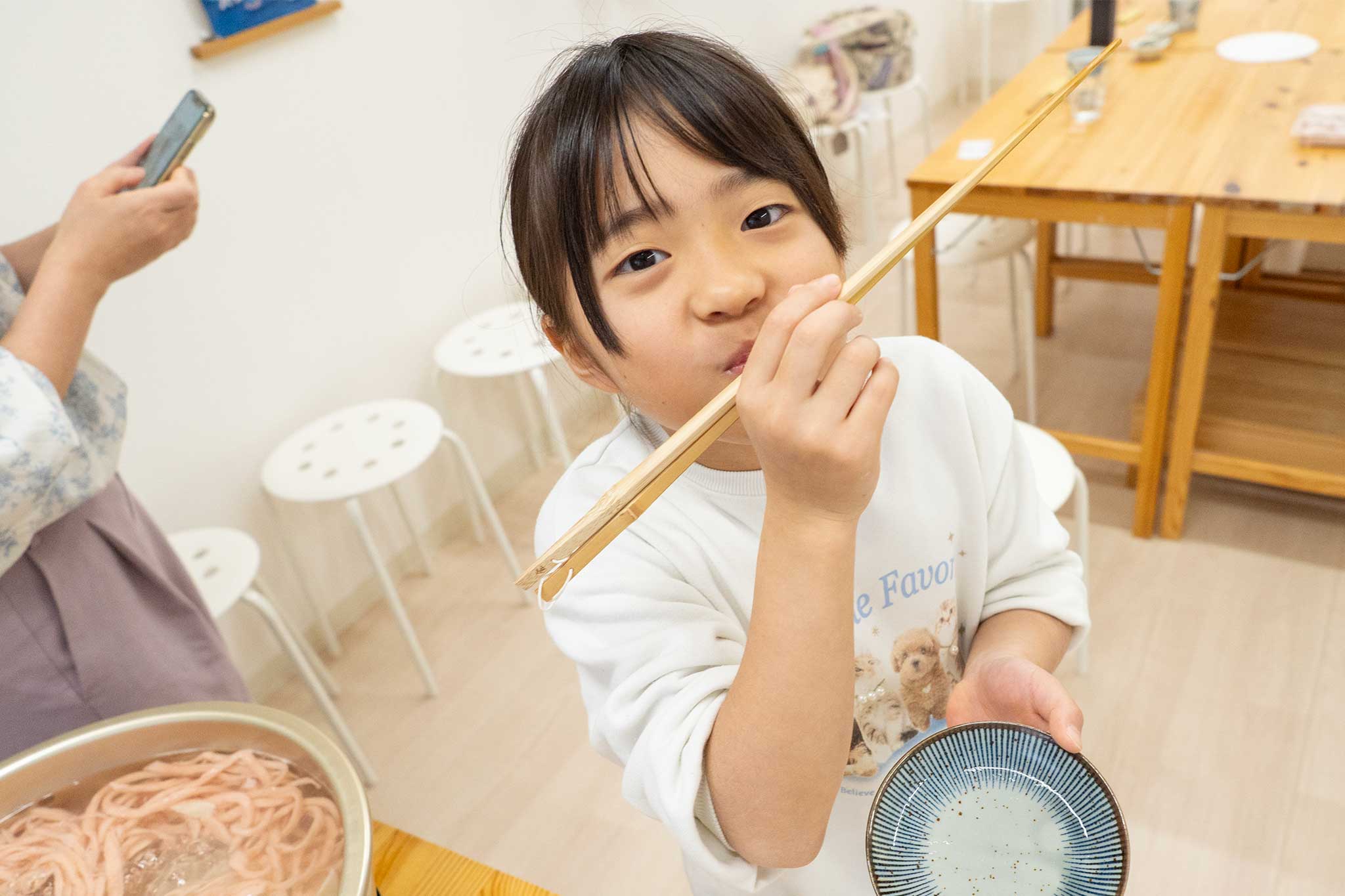 茹でたてのさくら色のうどんを鍋から菜箸でつまんで食べる女の子。つゆ椀を片手に持ち、ピンクの麺が鍋に見えている。 手打ちうどん作り体験教室 2026年3月 udon-class-sakura-udon-eating-from-pot-chopsticks.jpg – イリコスキー製麺所