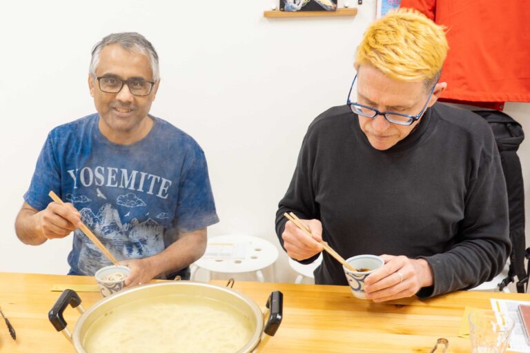 イリコスキーうどん作り体験教室で自作うどんを前に笑顔を見せる海外ゲスト2人。大鍋と薬味の並ぶテーブルを囲み、温かな雰囲気で体験を締めくくっている。 手打うどん作り体験教室 2026年1月 udon-class-tasting-smiling-guests-finish.jpg – イリコスキー製麺所