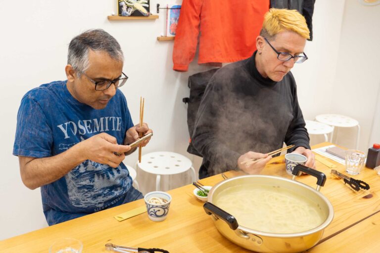 イリコスキーうどん作り体験教室で手打ちうどんを試食する海外ゲスト2人。湯気の立つ鍋を前に、1人はスマートフォンで撮影し、もう1人は箸でうどんをすくっている。 手打うどん作り体験教室 2026年1月 udon-class-tasting-steam-pot-two-guests.jpg – イリコスキー製麺所