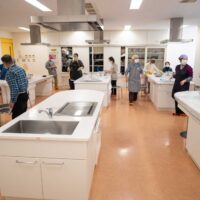 出張うどん体験教室の会場全景。調理実習室に参加者が集まり、各テーブルで作業の準備をしている。Participants
gathering at the cooking classroom for an outgoing udon-making workshop.