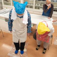 青いバンダナの男性がビニール袋に入れたうどん生地を足で踏んでいる。A man stepping on udon dough in a plastic bag
to knead it with his feet.