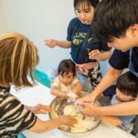 子どもと大人がひとつのボウルを囲んで、うどんの生地を混ぜている。Children and adults mixing udon dough together in one bowl. 出張手打ちうどん作り体験 2026年8月8日 20260808-1.jpg – イリコスキー製麺所