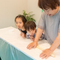 女性が麺棒でうどん生地をのばすのを、子ども2人がのぞき込んでいる。Two children watching closely as a woman rolls out udon dough with a rolling pin. 出張手打ちうどん作り体験 2026年8月8日 20260808-3.jpg – イリコスキー製麺所